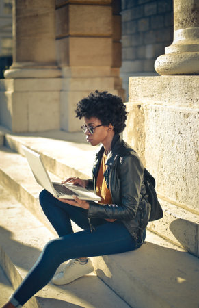 A vertical shot of a black female working on her laptop on the steps of an old buildingの写真素材
