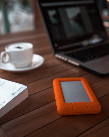 A selective focus shot of an external hard drive on a wooden table with a laptop computer and a cup...