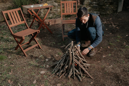 A young man preparing a pile of wood for fire with a table and chairs on the side - romantic dinnerの写真素材