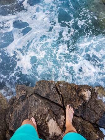 A top view of a male standing on a rocky seashore with hitting foam wavesの写真素材