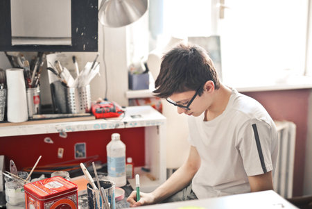 A boy with eyeglasses and a white t-shirt painting in his room