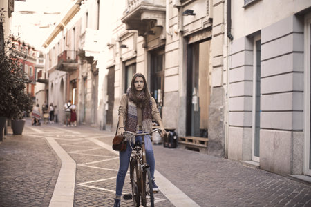A beautiful young lady with a scarf and jea riding a bike in a cozy streetの写真素材