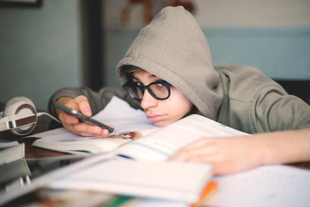 A tired boy with eyeglasses cheating and looking at his smartphone instead of doing homework