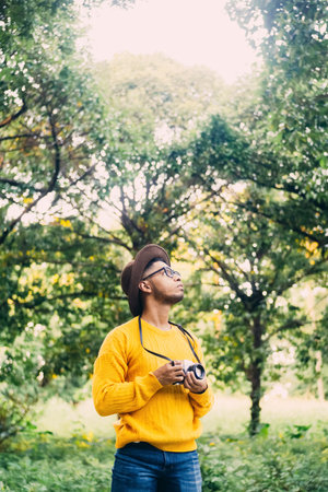 Young Latin American man in a green forest is wearing a hat, glasses and a yellow sweater holding...