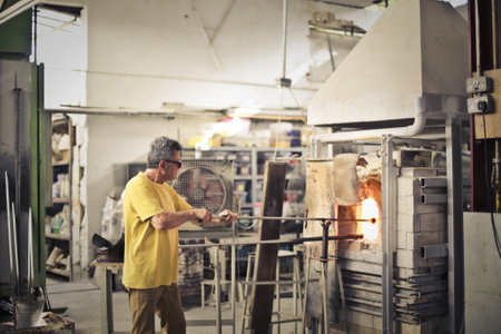 An old man shaping the glass with long tools in the oven at the glass factory