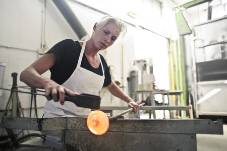 The blonde woman shaping the hot glass with tools in the glass factoryの写真素材