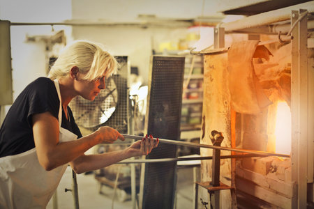 The blonde woman shaping the hot glass in the oven in the glass factoryの写真素材
