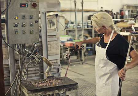 The blonde woman shaping the hot glass with tools in the glass factoryの写真素材