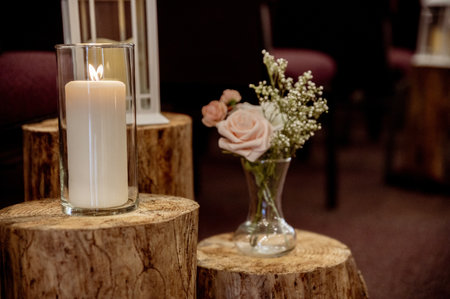 A selective focus shot of lit candle in a glass jar on a wooden table, in front of a vase of flowersの写真素材