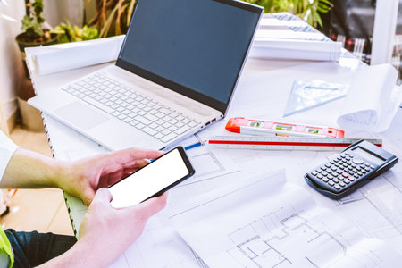 A male engineer hands using a mobile phone while working on blueprints at the desk