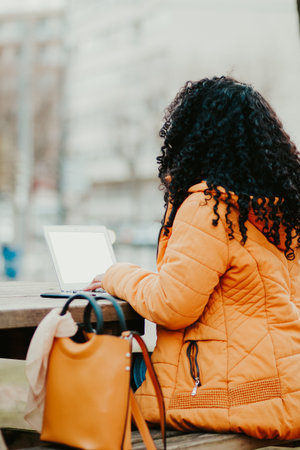A vertical shot of young curly woman sitting on a bench while using her laptop in the parkの写真素材