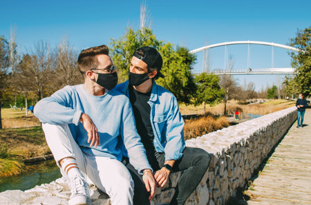 Picture of Lifestyle Gay Couple with Protective Black Face Mask and Sunglasses Together Looking at Each Other Sitting on Bridge Parkの写真素材