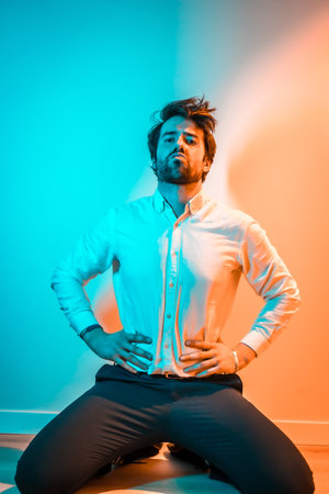 ACaucasian man on his knees in a white shirt and black pants, illuminated with an orange and blue...