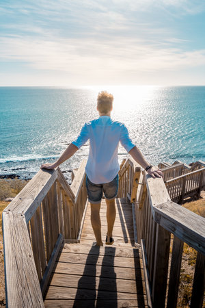 A vertical shot of a male from the back standing on the wooden stair and looking at the blue sea in Adelaide, Australiaの写真素材