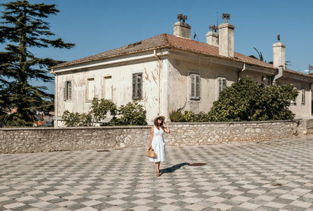 Full length portrait of young woman in white summer dress standing in middle of square in old town....