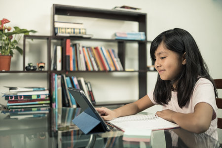 A happy Hispanic schoolgirl doing a homework using a notebook and tablet at home - online educationの写真素材