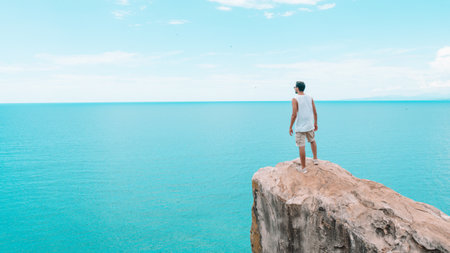 A young man enjoying the view of a calm sea from the top of a cliffの写真素材