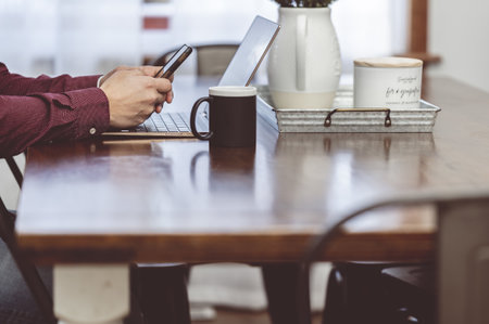 A man with a phone in his hands sitting next to a laptop and drinking a coffee in a cozy cafe