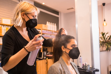 A hairdresser in a protective mask spraying client's hair with hairsprayの写真素材