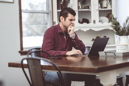 A shallow focus of a serious adult man working on a laptop at home with a blurry background