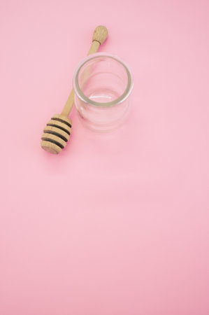 A top view closeup of a wooden honey dipper next to a glass container placed on a pink backgroundの写真素材