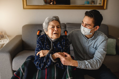 A young man with face mask talking to old sick grandmother in a wheelchair -family, home care conceptの写真素材