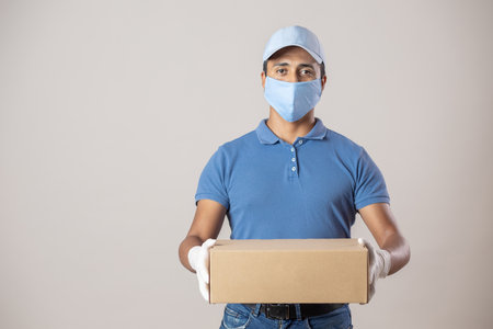 A delivery man employee in blue cap and t-shirt with face mask and gloves holding a boxの写真素材