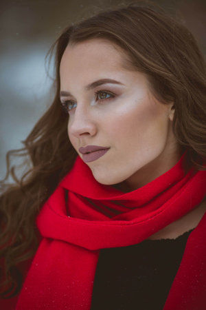 A portrait of a young brunette girl with perfect makeup wearing a stylish red scarf in a snowy forestの写真素材