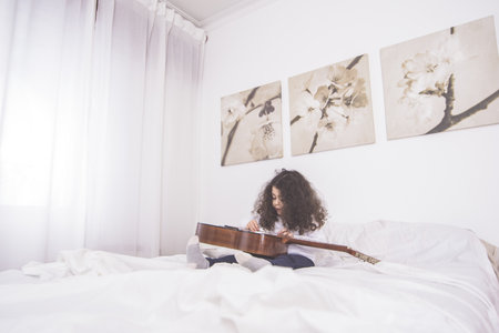 A closeup shot of a cute Caucasian little girl sitting in a bed with a guitar in her hands