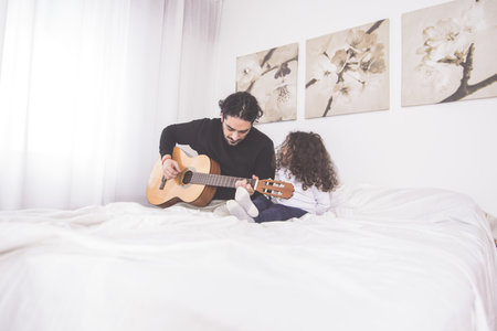 A father teaching his young daughter to play guitar sitting on a bedの写真素材