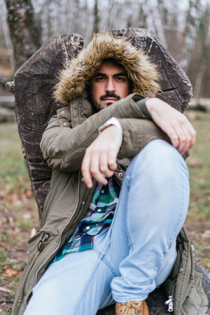 A vertical shot of a male in a winter jacket leaning against wood in a forest