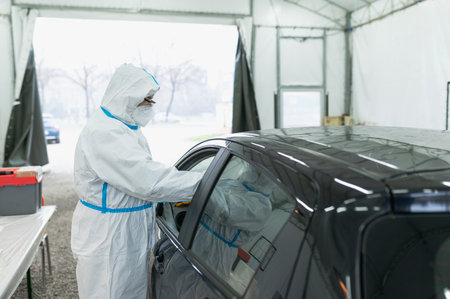 A nurse in a protective suit taking a sample from the driver inside the car in a drive-thru covid-19...