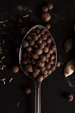 A vertical shot of a spoon with allspice berries on dark backgroun