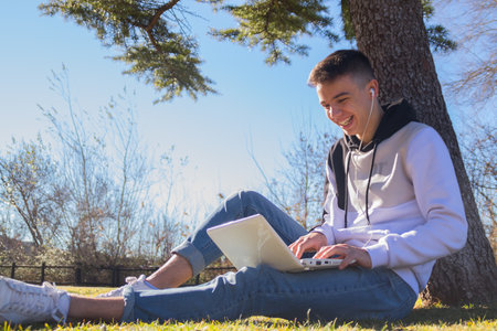 A cute Caucasian guy sitting on the grass, with his laptop on his lap and laughin