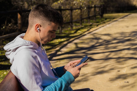 A cute Caucasian boy sitting on the bench with his phone and headphones in the parkの写真素材