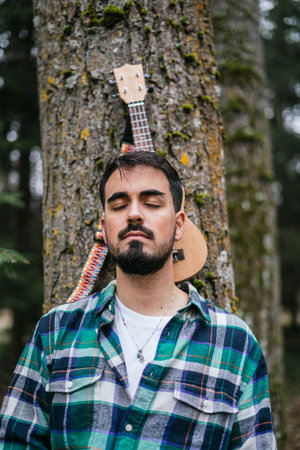 A bearded Hispanic man wearing flannel with eyes close while leaning on ukulele against a treeの写真素材