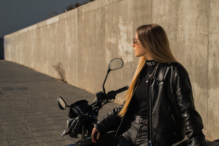 A pretty Caucasian lady wearing a leather jacket with an all-black outfit next to her motorbike