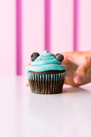 A female hand about to take a chocolate cupcake topped with blue cream and blueberriesの写真素材