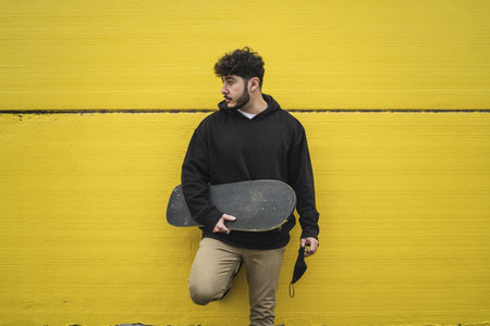 A cool European male skateboarder leaning on a vibrant yellow wall