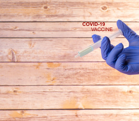 A hand in a medical glove holding a syringe with the text "Covid-19 vaccine" on wooden backgroundの写真素材