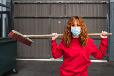 A female worker wearing a mask posing with a large broom behind her backの写真素材
