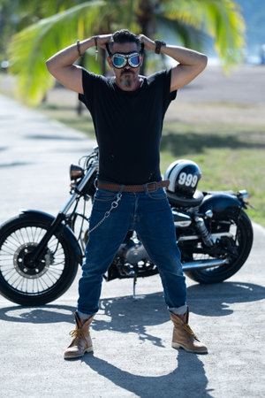 A strong male with a black shirt and eyeglasses standing in front of his motorbikeの写真素材
