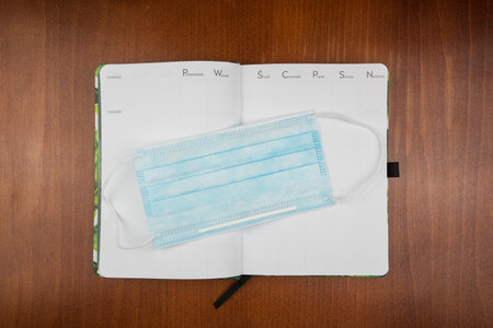 A top view of an open empty weekly dairy planner with a face mask on an office wooden desk