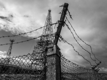 A grayscale shot of a power transmission tower behind barbed wiresの写真素材
