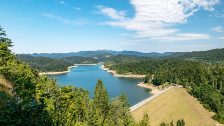 A landscape of a lake surrounded by hills covered in greenery under the sunlight and a blue skyの写真素材