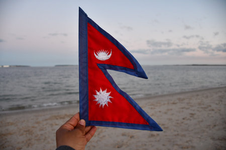 A selective focus of a hand holding the flag of Nepal on blurred sea backgroundの写真素材