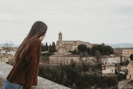 A young female in a brown sweater standing on a bridge and enjoying beautiful view of the cityscapeの写真素材