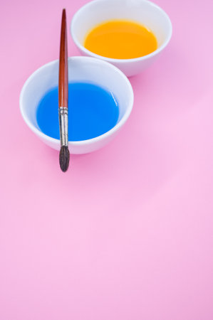 A vertical shot of blue and yellow liquid watercolor paints on a bowl and paintbrush isolated on pink backgroundの写真素材