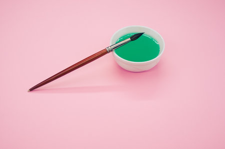 A high angle shot of green liquid watercolor paint on a bowl and paintbrush isolated on pink...
