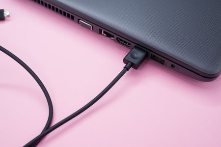 A closeup of a black laptop with a cable connected on pastel pink surfaceの写真素材